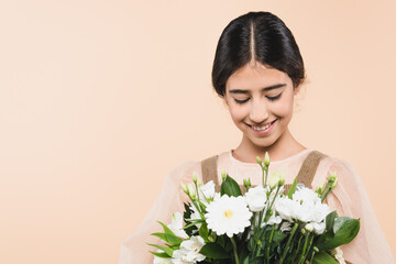 Cheerful hispanic girl looking at flowers isolated on beige
