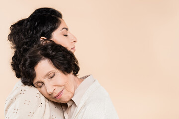 Senior woman with closed eyes standing near hispanic daughter isolated on beige, two generations of women