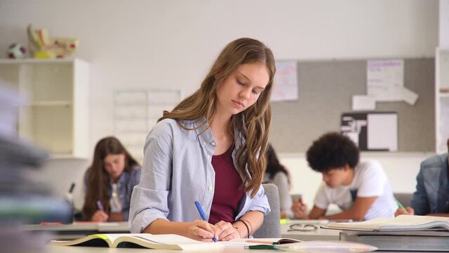 Portrait of smiling high school student with classmates in background writing notes in the classroom. Happy casual girl sitting at desk in class while looking at camera. 
