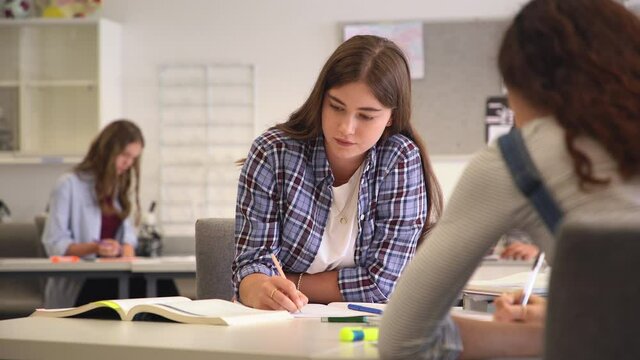 University student smiling while studying for exams in classroom. Pretty girl sitting in classroom full of students during class. Portrait of happy young woman writing notes and looking at camera.