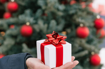 Man holds in his hand gift box with red ribbon on the background of a Christmas tree and Christmas balls. Selective focus. Blured background