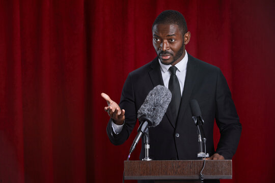 Waist up portrait of African-American man speaking to microphone standing at podium on stage against red curtain, copy space