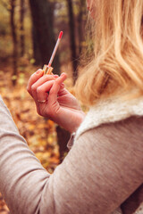 A young woman in a beige cozy, knitted sweater holds a lip gloss in her hands. Woman applying makeup on a background of golden autumn foliage. Express makeup