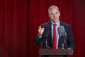 Waist up portrait of mature man speaking to microphone standing at podium on stage against red curtain, copy space - Powered by Adobe