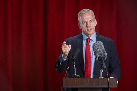 Waist up portrait of mature man speaking to microphone standing at podium on stage against red curtain, copy space