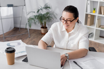 Obraz na płótnie Canvas Smiling plus size hispanic businesswoman typing on laptop at workplace with papers and documents on blurred background