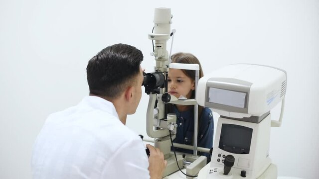 Young oculist in white coat using special device to check little girl's eye.