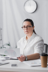 Serious plus size hispanic businesswoman looking at camera while holding paper at workplace on blurred foreground