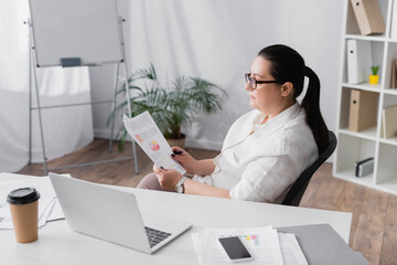 Plus size hispanic businesswoman looking at papers with graphs while sitting at workplace with devices on blurred background