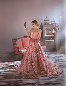 beautiful woman in sexy long medieval pink dress, spring flowers on skirt, looks at herself. Holds in hand vintage mirror. Adult girl queen with big breasts in corset, open chest. Elegant bun hair