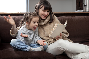 Young mother with her daughter are reading a book at home while sitting on the couch.