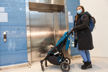 Mother wearing face mask waiting elevator with children pram in hall of shopping mall