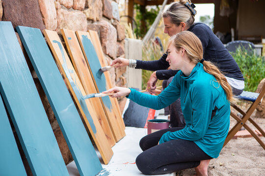 Teenage girl and her mother painting wooden shelves blue on a terrace