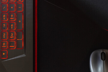 Laptop keyboard with red backlight, computer mouse on a black mouse pad.