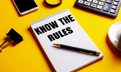 On a yellow background, a notebook with the text KNOW THE RULES, coffee, calculator, telephone and a clip for papers. Business concept