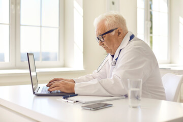 Aged professional doctor in uniform sitting at laptop and communicating with patient online