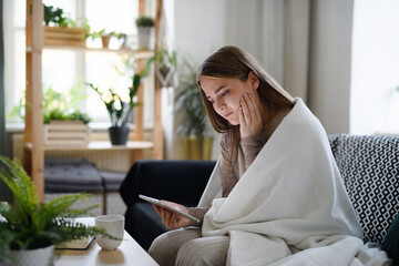 Worried young woman with blanket using tablet at home, coronavirus concept.