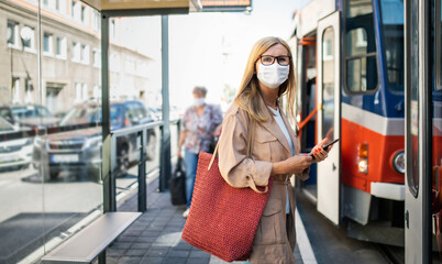 Senior woman with smartphone on bus stop outdoors in city or town, coronavirus concept.