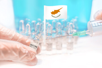 Hands holding a vaccine vial and a syringe with rows of the same capsules and a flag of Cyprus in the background, illustrating plans for global vaccination against Covid-19 (SARS-CoV-2).