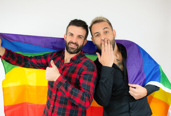 Happy gay couple wrapped with the homosexual flag with thumb up over white background.