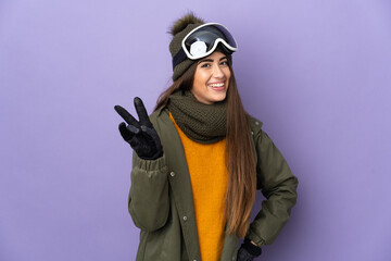 Skier caucasian girl with snowboarding glasses isolated on purple background smiling and showing victory sign