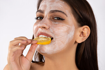 A girl with cream on her face holding slice of lemon in her mouth. White background. Spa and beauty