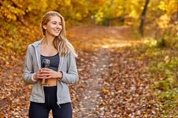 athletic woman holding a clean bottle of water in the autumn forest, blonde female woman of European caucasian appearance is engaged in healthy lifestyle, wellbeing