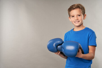 Joyful boy with boxing gloves standing against gray background
