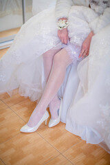 Fototapeta na wymiar portrait of the bride in a white wedding dress in white heel shoes wears a garter on her leg, the bride is holding her hands for the garter.