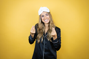 Young pretty woman wearing sleep mask and pajamas over isolated yellow background doing money gesture with hands, asking for salary payment, millionaire business