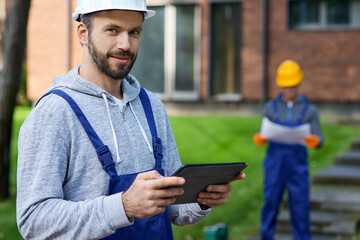 Portrait of young male builder smiling at camera while holding digital tablet pc with coworker working in the background at cottage construction site