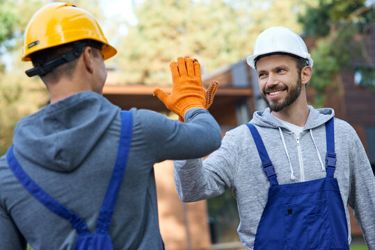 Leave It To The Professionals. Two Positive Young Male Engineers In Hard Hats Smiling At Each Other, Giving High Five While Working On Cottage Construction Site Outdoors