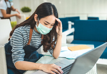 Upset cafe owner holding paper near calculator, laptop and document on table.Asian business woman with face mask having a headache while sitting in a cafe.