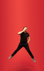 Fototapeta na wymiar Talking phone. Full length portrait of young high jumping man gesturing isolated on red studio background. Attractive male caucasian model. Copyspace. Human emotions, facial expression concept.