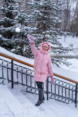 Young woman in a pink down jacket in a city park standing on the stairs throws snowballs