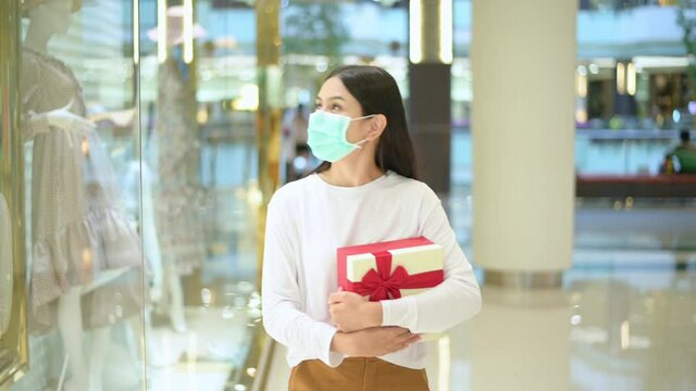 woman wearing protective mask holding a gift box in shopping mall, shopping under Covid-19 pandemic, thanksgiving and Christmas concept.
