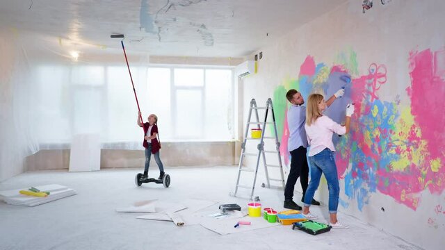 modern kid paints the ceiling with a roller on a hoverboard in a new apartment, little girl helps parents during the renovation