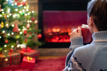 Woman with cup of hot cocoa and marshmallow sitting and warming at winter evening near fireplace flame and  christmas tree.