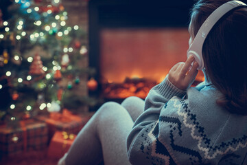 Fototapeta na wymiar Woman in headphones sitting and warming at winter evening near fireplace flame and christmas tree.