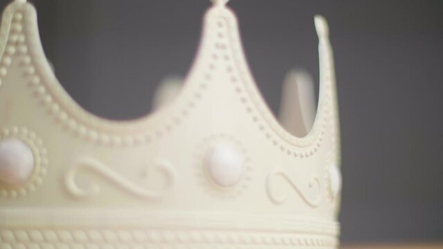 Close up of a beige crown spinning on a pedestal with ornaments and white pearl jewels with peaks jagged out of plastic. With a neutral grey background. Shallow depth of field. Shot in 4K.	