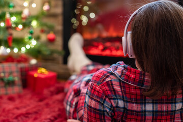 Woman in pajama in headphones lying, relaxing and warming at winter evening near fireplace flame christmas tree.