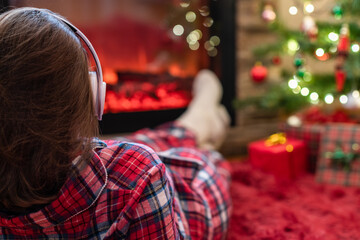 Woman in pajama in headphones lying listening music and warming at winter evening near fireplace flame christmas tree.