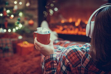 Woman in pajama in headphones with cup of hot cocoa and marshmallow lying and warming at winter...