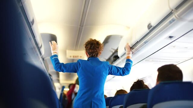 Masked stewardess in gloves controls closes boxes with hand luggage on aircraft board. Baggage rules during social distancing, reduce travel in cabin.
