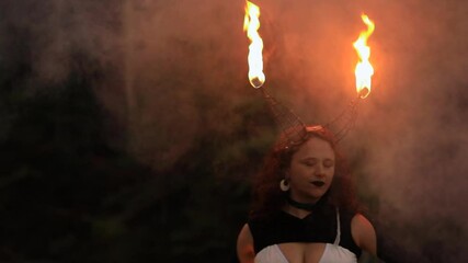 Portrait of beautiful young female wearing metal horn with flame using spinning fire with smoke in forest during outdoor fire jamming