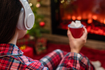 Woman in pajama in headphones with cup of hot cocoa and marshmallow sitting and warming at winter...