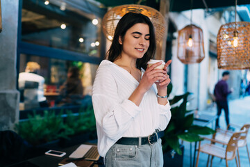 Carefree hipster girl dressed in casual white shirt looking at disposable cup with caffeine beverage and smiling during rest time in city, happy Caucasian woman enjoying coffee break outdoors