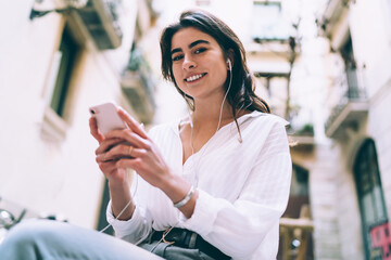 Half length portrait of cheerful hipster girl with smartphone technology in hands smiling at camera during music pastime, happy female teenager in headphones listening positive online radio