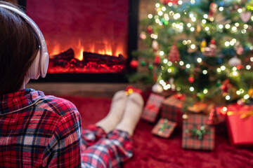 Obraz na płótnie Canvas Woman in pajama in headphones sitting and warming at winter evening near fireplace flame and christmas tree.