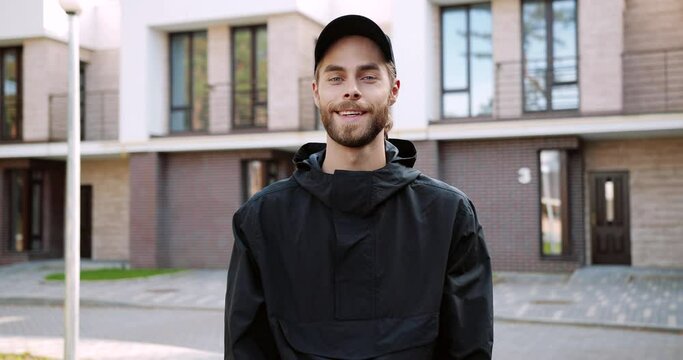 Portrait of Caucasian young joyful guy in cap standing on street in city, looking at camera and smiling. Handsome happy delivery man in good mood at work. Occupation concept